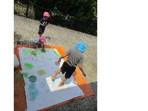 写真：外のレジャーシートの上に大きな用紙を敷き、手のひらや足の裏に絵の具を塗って、お絵かきを楽しむ子どもたち。