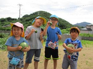 写真：4人の子どもたちがそれぞれ畑の収穫した小玉スイカやナス・ピーマンを持っています。