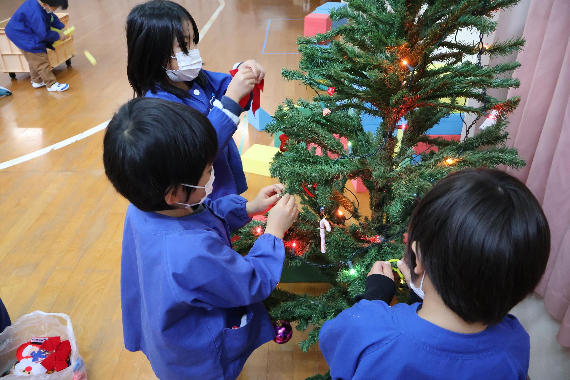 写真：クリスマスツリーにリボンや装飾の飾りつけをする2人の男の子と一人の女の子