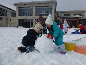 写真：2歳児の男の子2人と女の子1人が大きな雪玉を転がしています。