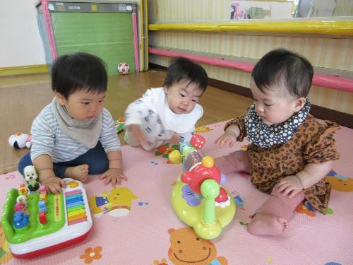 写真：ひよこ組の保育室の様子。まわすと音のなるおもちゃで遊ぶ0歳児。その横にいる2人の子も興味津々でのぞきこんでいます。
