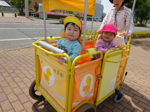 写真：お散歩の様子。立ち乗りバギーに乗って、笑顔をみせる1歳児の新入児二人