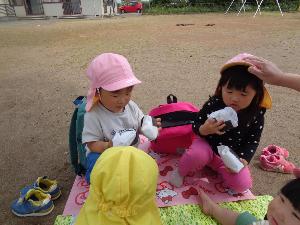 写真：2歳児の子どもたちが園庭でレジャーシートを広げ、手には紙で作ったおにぎりを持ち、遠足ごっこをしています
