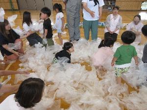 写真：たくさんのビニール袋の中で園児たちが座ったり、ビニール袋を放り投げたりして遊んでいます。その周りを保護者が囲んでいます