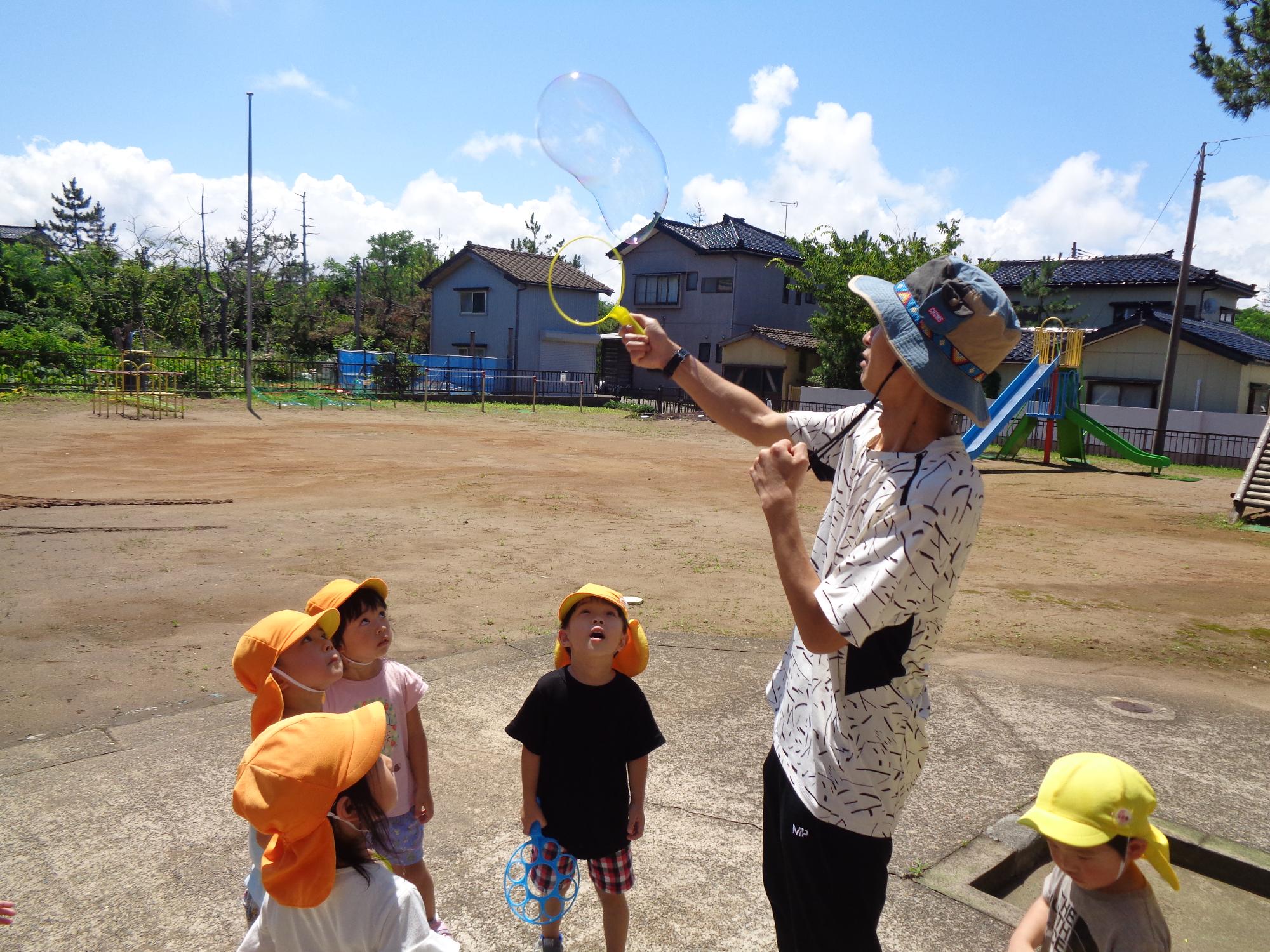 写真：年少児4人が保育者の作っている大きなしゃぼん玉を見上げている様子。