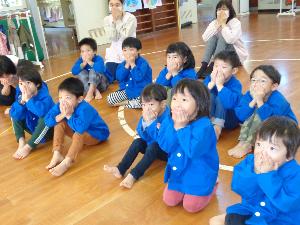 写真：お遊戯室に集まった園児たちが両手で口を押えてアライグマのポーズをしているところ