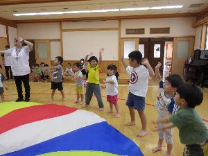 写真：年長組に教えてもらいながら、運動会のバルーン競技を楽しむ2歳児たち。料手を大きく振って笑い合っています