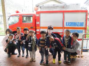 防火の法被を着た7人の子ども達と担任と消防士が消防車の前で記念撮影