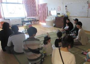 写真：保育室で親子で座って映画を楽しんでいるところ。保護者の膝の上に座る子、横に並んで座る子とそれぞれでゆっくり過ごしています