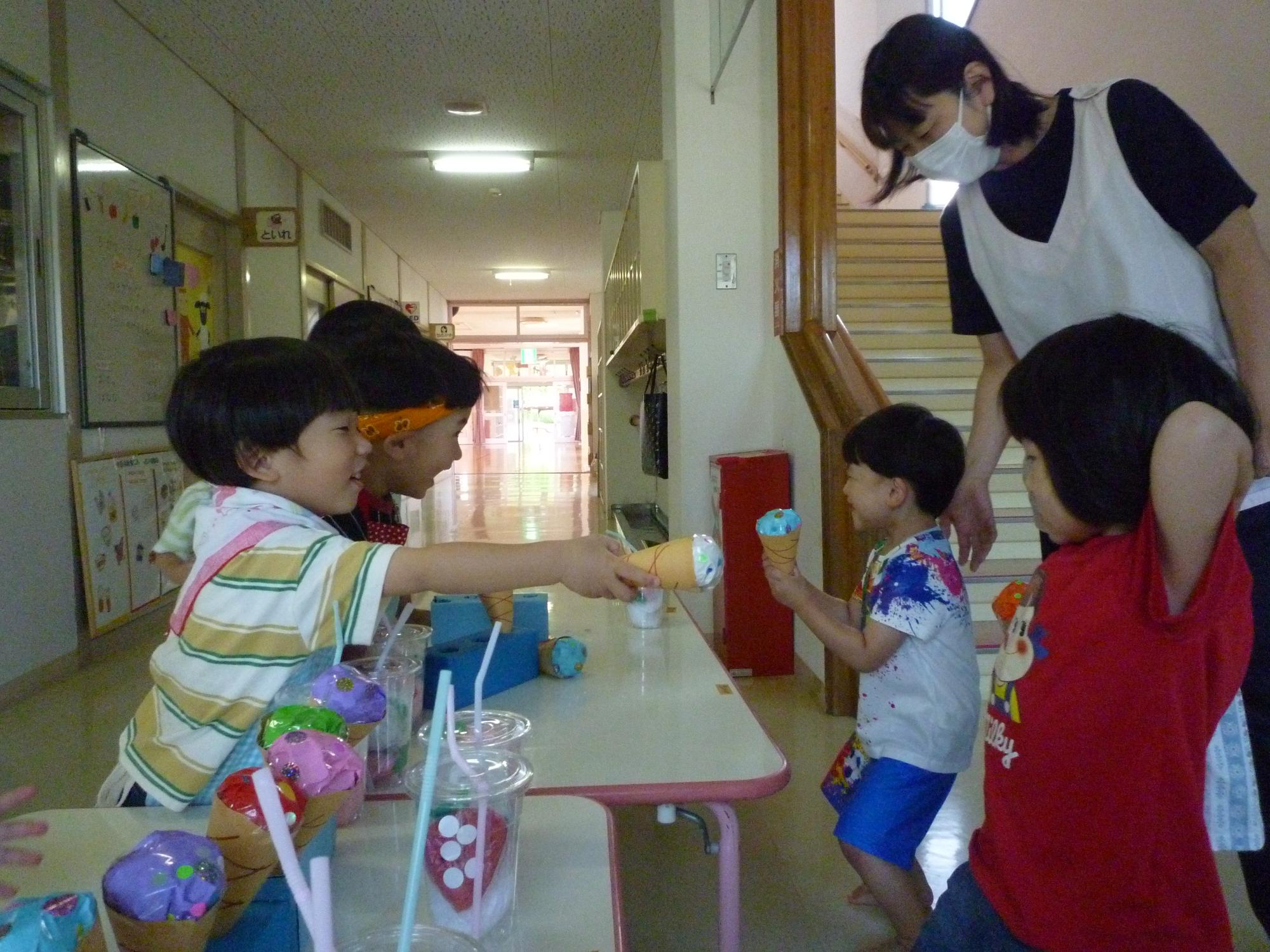 写真：えんま市ごっこの様子。2歳児の子どもたちがアイス屋さんになり、色画用紙で作ったコーンアイスを3歳児クラスの子どもに渡しているところ