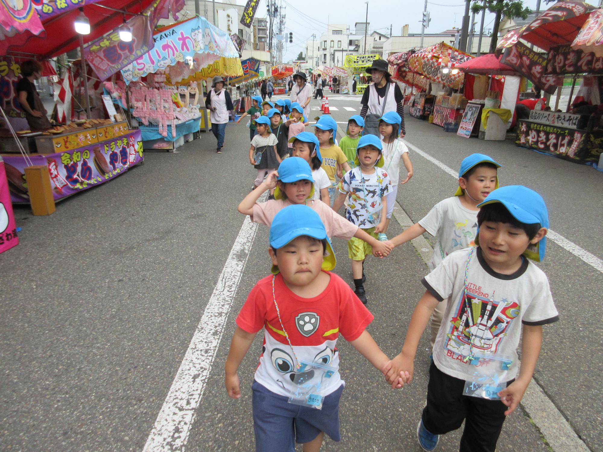 写真：両脇に露店が並ぶ通りを、2人1組で手をつないで歩く5歳児クラスの子どもたち