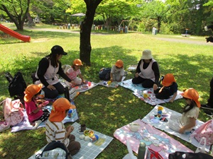 写真：木陰のある芝生の上でレジャーシートを広げ、ぱんだ組の子どもたちが輪になってでお弁当を食べています