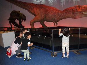 写真：赤くライトアップされた恐竜の大型模型を見ている園児たち。時折、動いたり吠えたりする恐竜に少し怖がっている様子です。