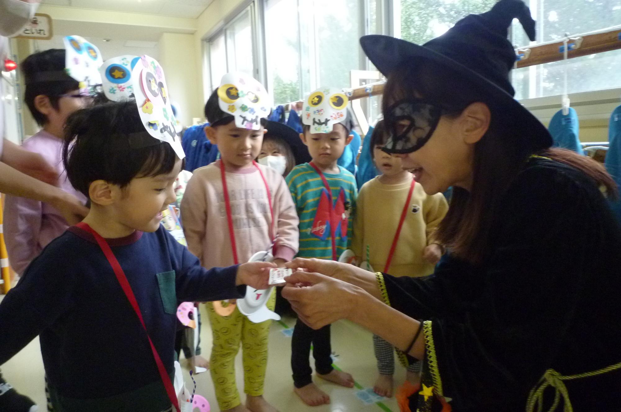 写真：ミッションをクリアした子どもが、魔女に変装した職員からご褒美のチケットをもらっています。その後ろには同じチームの子どもたちが並んでいます。