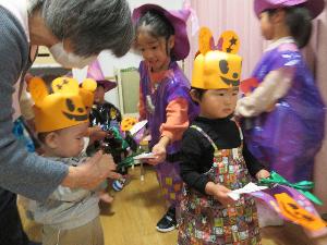 写真：オバケに扮した5歳児と4歳児が待つ保育室の中に、1歳児と0歳児の子どもたちがやって来た場面。オバケたちからプレゼントされた紙皿と折り紙で作ったハロウィンのリース飾りを持っています。