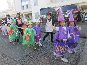 写真：カラーポリ袋や色画用紙で魔女などのオバケに扮装した5歳児を中心とした8人ほどが、2列に並んで歩きだそうとしているところ。このあと、地域5カ所を回ります