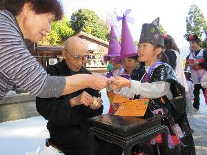 写真：周廣院の住職が、訪れた子どもたちのカードにシールを貼ろうとしています。奥さまは工作したお菓子を子どもたちに手渡しています。