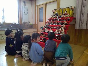 写真：7段飾りの雛人形の前で正座しながら、うれしいひなまつりの歌を歌う子どもたち。6人のかわいらしい後ろ姿が並んでいます。