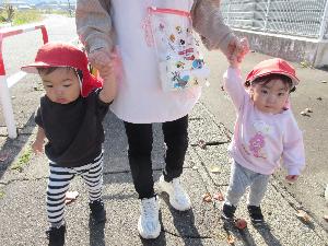 写真：保育士と手をつないで、1歳児2人が歩道をあるいているところ