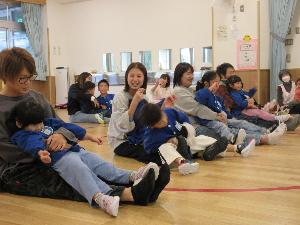 写真：広い遊戯室で、膝を伸ばして座る保護者の足の上に、子どもたちが座っています。横や縦に足を揺らしたりして、親子とも楽しそうにしています。