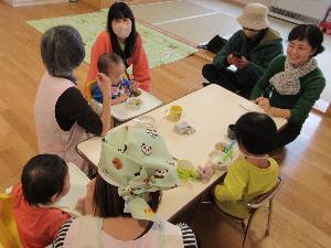 写真：給食参観と試食会の様子。0・1歳児の教室で子どもたちが給食を食べています。その横に保護者が座り、園での食事の様子や家庭での食事に関する困りごとなどを職員と話しています。