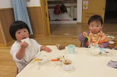 0・1歳児2人が遊戯室での会食に参加し、できあがったおでんを食べている写真