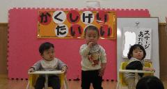 写真：3人の園児が手遊び歌を披露しています。真ん中の子どもが手を口に運び、食べるしぐさをしています。