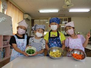 写真：エプロンとマスクを着用した年長・年中児4人が、自分たちが切った野菜の入ったボウルを持っています。ボウルの中にはピーマン、ジャガイモ、ニンジンが見えます。