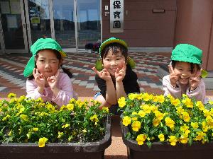 写真：玄関前に置かれたプランターに咲く黄色い小花。その後ろに3人の女の子が座りポーズを決めています