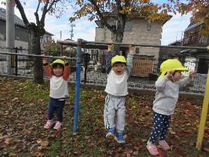 写真：2歳児の3人が秋の公園で鉄棒につかまって喜んでいるところ
