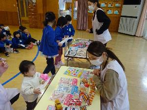 写真：抽選に当たったはがきを手に持ち、玩具が並べてある机の前に立つ0歳女児