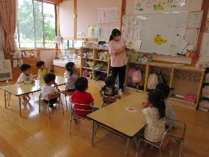 写真：保育者が絵本を読み聞かせています。子どもたちは椅子に座って、お話しに聞き入っています。