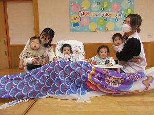 写真：保育士に抱っこされたひよこ組の0・1歳児の子どもたちがおおきなこいのぼりと一緒に写真を撮りました