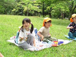 写真：レジャーシートを敷いた芝生の上で、お弁当を食べながら笑顔を浮かべる子どもたち