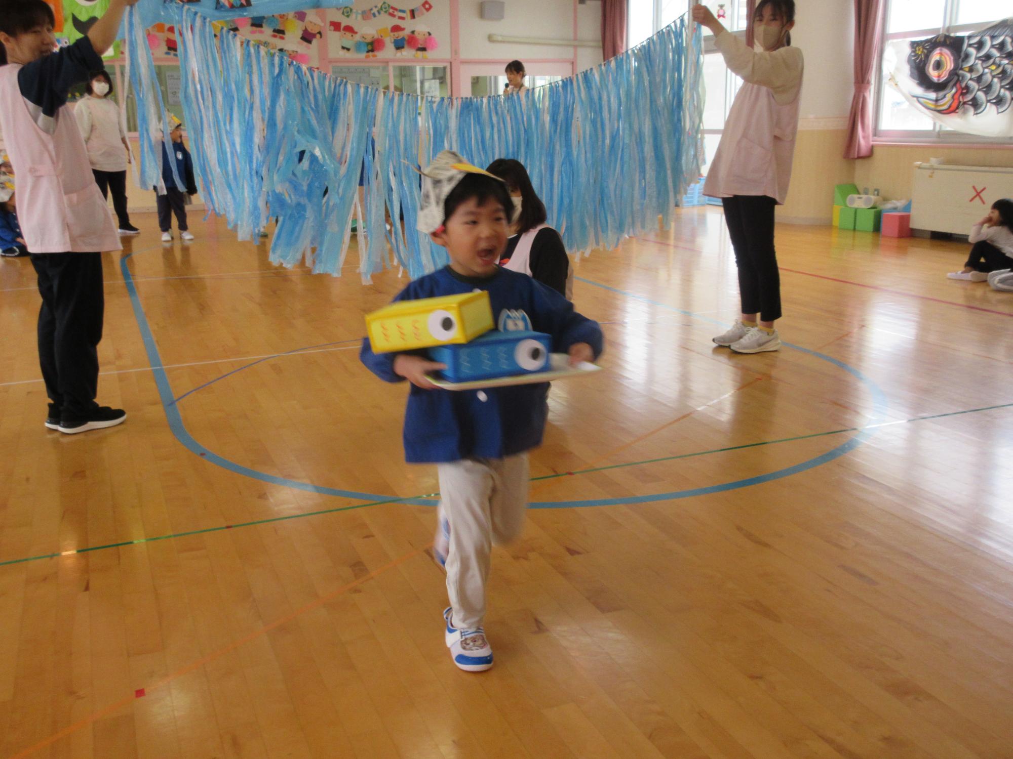 写真：らいおん組の子どもが重ねたこいのぼりをトレーに載せて、落とさないように運ぶリレーをしています
