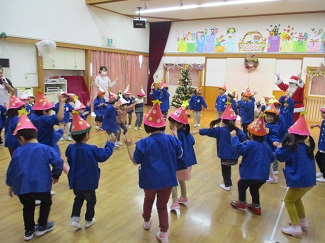 写真：遊戯室で子どもたちがサンタさんと輪になってフォークダンスを踊っている様子