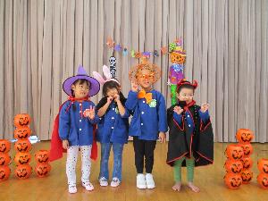 写真：オバケかぼちゃの装飾の前で、仮装しポーズを取る4人の3・4歳児