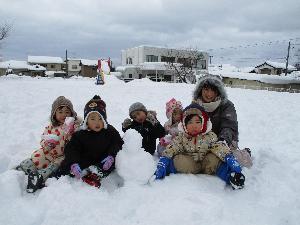 写真：雪上で雪だるまと一緒に座ってポーズをとる2歳児数人と保育士