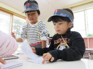 写真：5歳児2人が郵便マークのついた帽子を被り、郵便ごっこはがきにハンコを押しています。
