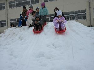写真：5歳児1名と3歳児1名が一人ずつそりに乗って雪山を滑っています。後ろには順番待ちの列ができています