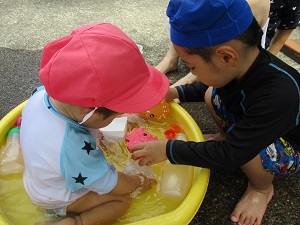 写真：たらいで水遊びをする1歳児男児とその前にしゃがみおもちゃで一緒に遊ぶ3歳児男児