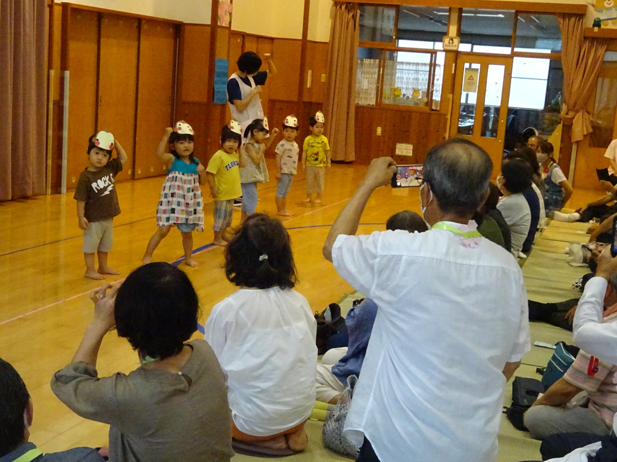 写真：お招き会で踊る2歳児。その様子をカメラに収めようと、おじいちゃんおばあちゃんがカメラを構えています。