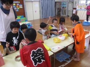 写真：お店屋さんごっこの食べ物屋さんで、買い物をする4歳児5人とお店屋さん役の5歳児2人が写っています。お店屋さん役の年長児はハッピを着ています