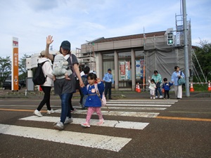 写真：保護者と一緒に信号機付きの横断歩道を、しっかり手をあげて渡る園児たち