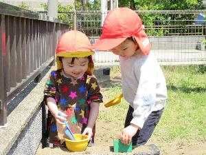 写真：2歳児の子どもたちが園庭の砂場でおままごとをして遊んでいます。