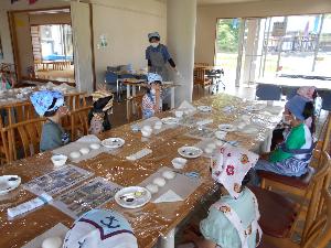 写真：年長児がこども大学のプログラムで米粉のパン作りの説明を聞いています。子どもたちの前にはそれぞれに、パン生地やトッピングが並べられています。