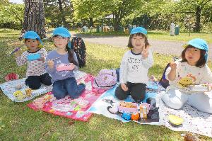 写真：4人の女児が芝生の上に広げたシートの上に座りお弁当を食べています。