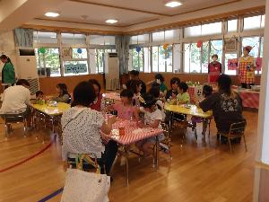 写真：遊戯室に並んだテーブルについて、子どもたちとおうちの方一緒に手作りおやつを食べています
