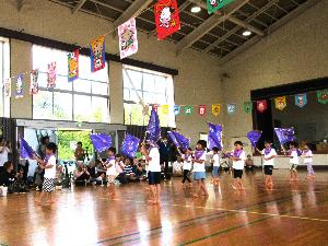 写真：子どもたちが手作りした紫色のカラーポリ袋製のフラッグを持ち、横二列になって曲に合わせて演技をしています。会場の上部には動物柄の万国旗が飾られています