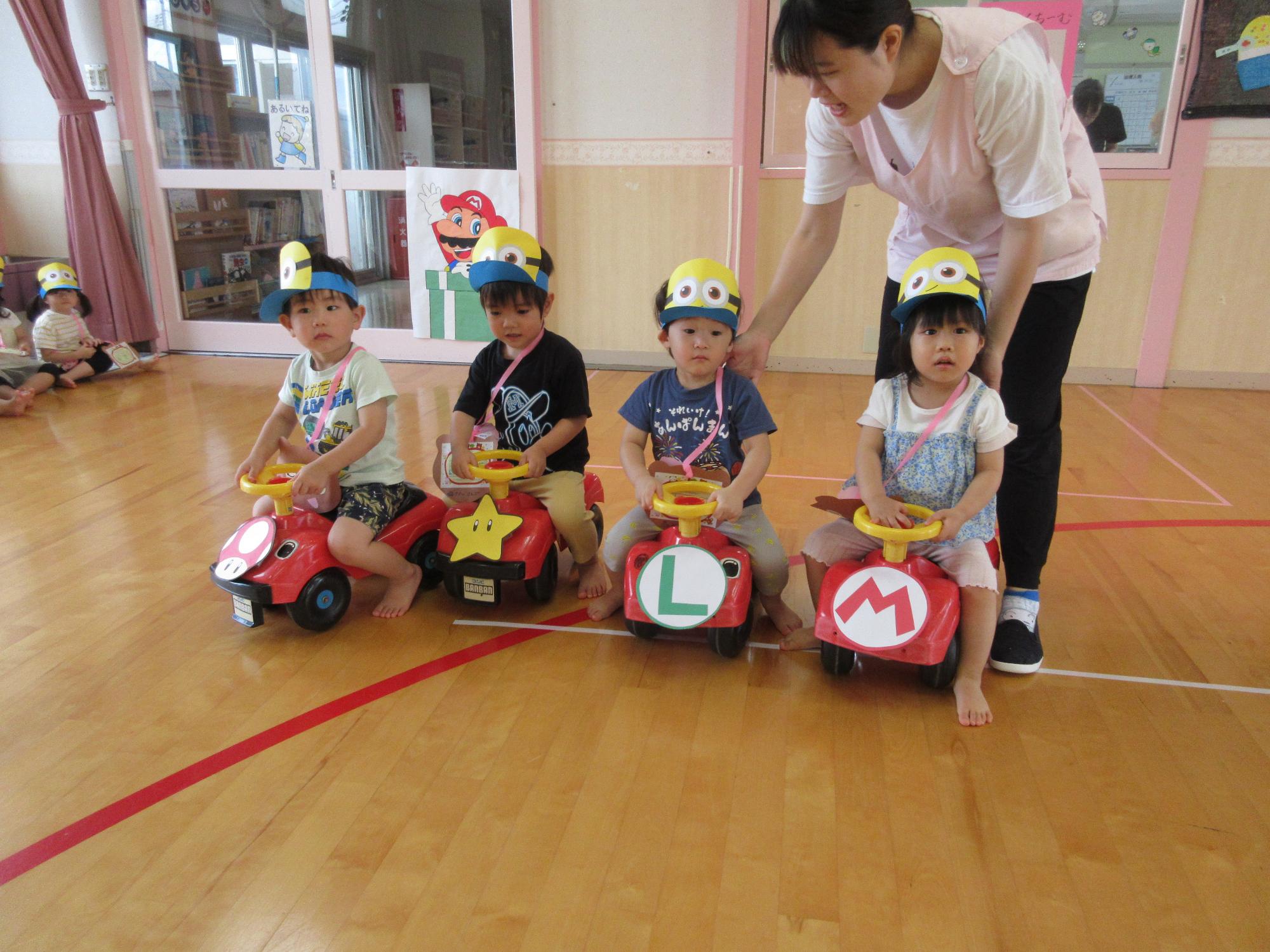 写真：マリオの絵を貼った乗用おもちゃに乗る2歳児4人の子どもたち。ミニオン（一つ目の黄色いキャラクター）の帽子をかぶっています
